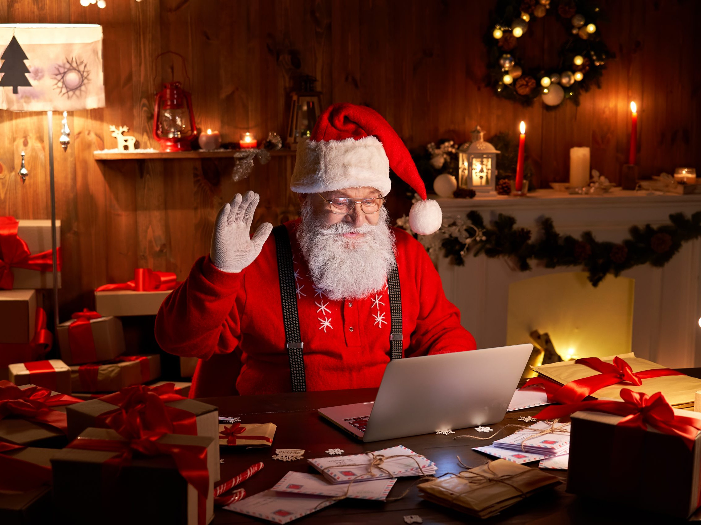 Fröhlicher Weihnachtsmann winkt im Videoanruf einem Kind zu, das an einem Online Weihnachtsfeier Quiz teilnimmt, während er spät am Heiligen Abend zu Hause am Laptop sitzt und Geschenke präsentiert.